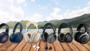 An assortment of the best noise-cancelling headphones and earbuds displayed on a wooden table with a scenic lake and lush forested mountains in the background. From left to right: Bose QuietComfort 45 in black, Sony WH-1000XM5 in blue, Apple AirPods Max in silver, Shure AONIC 50 Gen 2 in black, Soundcore Life Q30 by Anker in black, and Beyerdynamic Amiron Wireless headphones in black and silver. Below the over-ear models are Apple AirPods Pro 2 earbuds in white and Bose QuietComfort Earbuds in black, showcasing a range of premium audio options for audiophiles and neurodiverse travelers.