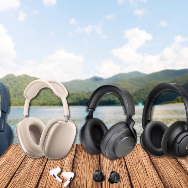 An assortment of the best noise-cancelling headphones and earbuds displayed on a wooden table with a scenic lake and lush forested mountains in the background. From left to right: Bose QuietComfort 45 in black, Sony WH-1000XM5 in blue, Apple AirPods Max in silver, Shure AONIC 50 Gen 2 in black, Soundcore Life Q30 by Anker in black, and Beyerdynamic Amiron Wireless headphones in black and silver. Below the over-ear models are Apple AirPods Pro 2 earbuds in white and Bose QuietComfort Earbuds in black, showcasing a range of premium audio options for audiophiles and neurodiverse travelers.