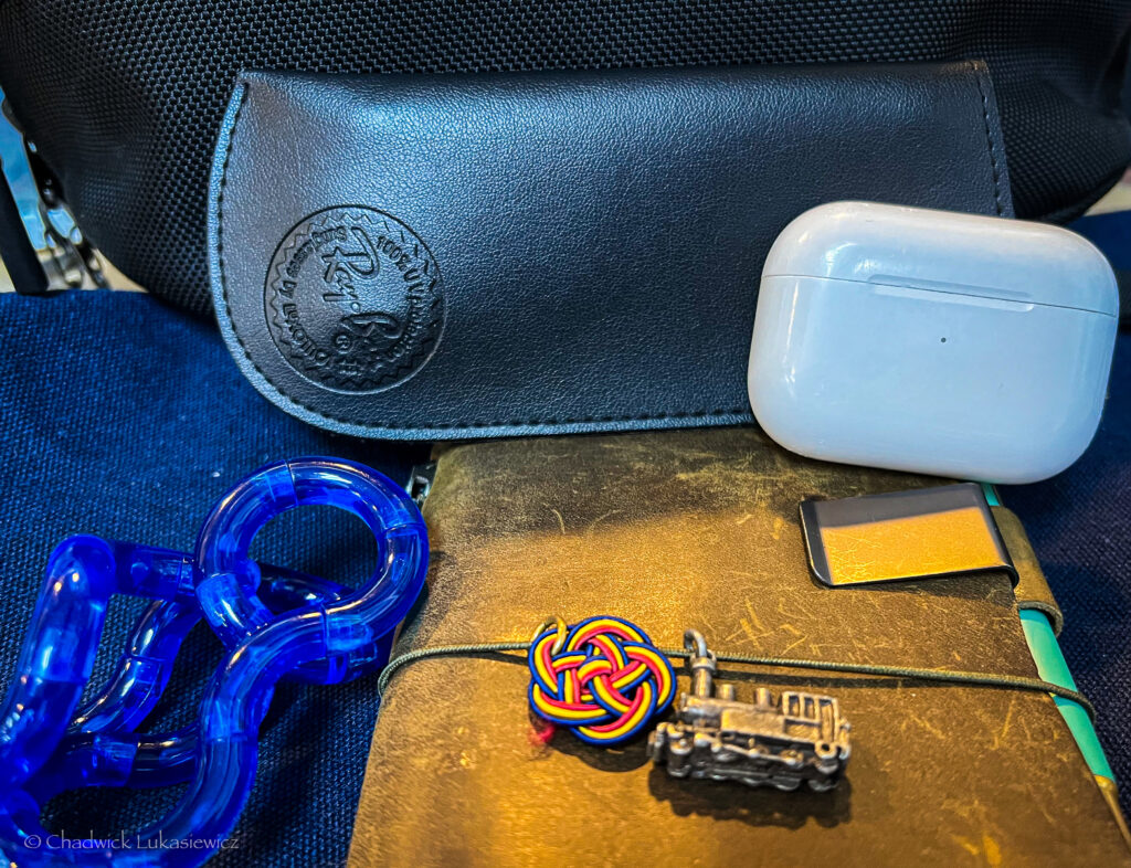 A close-up shot of personal travel essentials laid out on a surface. The items include a pair of white AirPods Pro, a leather-bound Traveler’s Notebook adorned with a colorful knot and a small train charm, a black sunglasses case embossed with a logo, and a blue fidget toy. The arrangement is casual, with the black bag in the background partially visible. The mix of textures—leather, plastic, and metal—creates a dynamic composition