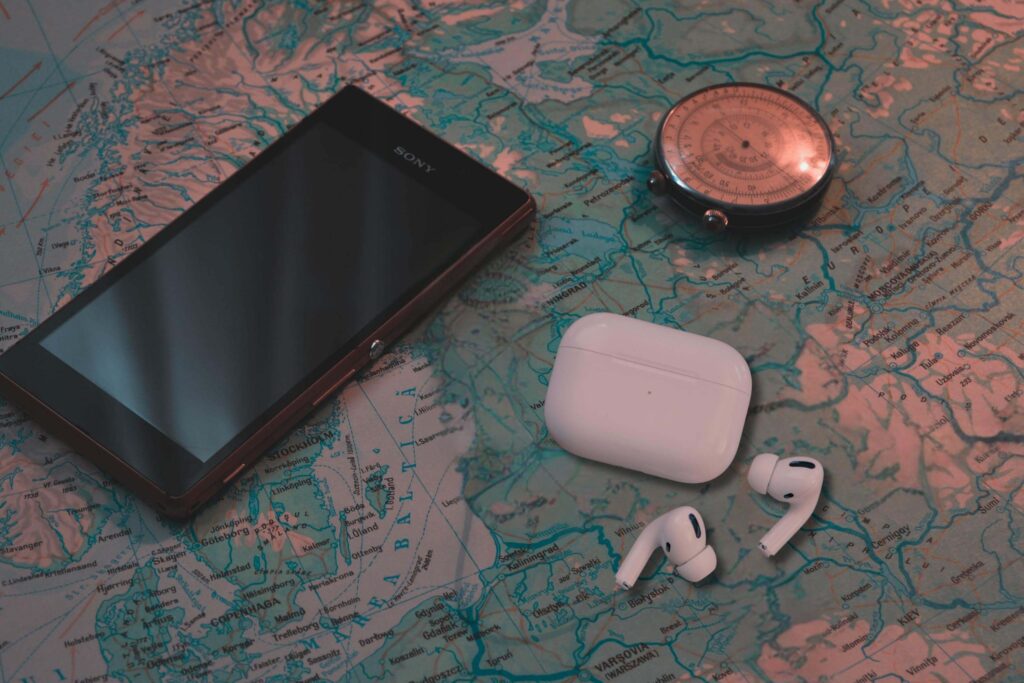 A pair of Apple AirPods Pro earbuds and their white charging case are placed on a detailed world map. To the left, a Sony smartphone with a reflective screen lies partially open. Above the AirPods, a vintage compass with a metallic finish rests on the map, creating a travel-themed arrangement.