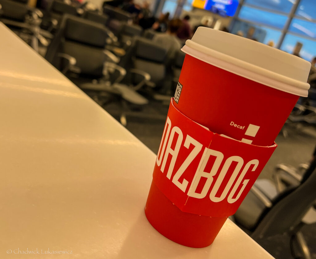 A red Dazbog coffee cup with a white lid sits on a smooth counter in an airport lounge. The cup sleeve prominently displays the bold white Dazbog logo. The background is slightly blurred, showing rows of gray airport seating and travelers waiting, with soft lighting from the terminal’s windows and screens visible in the distance. The image conveys a moment of quiet at the airport, perhaps while waiting for a flight.