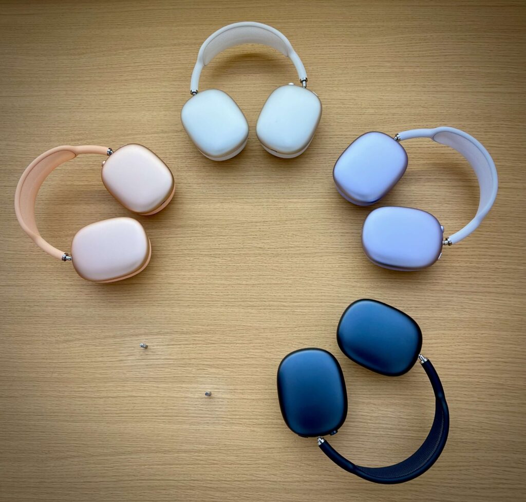 Four pairs of Apple AirPods Max headphones in various colors—pink, white, purple, and black—arranged in a circular pattern on a light wooden surface.