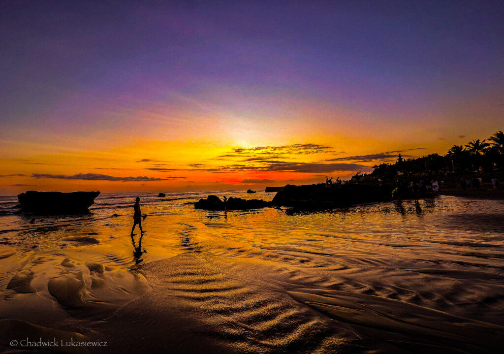 A stunning sunset on Canggu Beach, Bali, with the sky illuminated in shades of orange, yellow, and purple. Silhouettes of people can be seen walking along the wet, reflective sand, with a lone figure in the foreground. Large rocks are scattered in the shallow water, and a group of people gather in the distance near the shoreline, enjoying the view as palm trees and distant buildings frame the horizon.