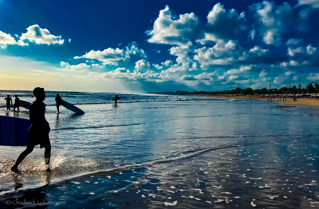 Surfers at Legian Beach, Bali, walking along the shoreline with their boards at sunset. The sky is a vivid blue with fluffy white clouds, and the ocean waves roll in softly. The silhouette of a surfer walking in the foreground creates a dynamic scene against the reflection of the sky on the wet sand.