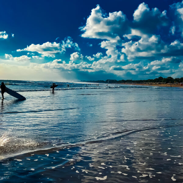 Surfers at Legian Beach, Bali, walking along the shoreline with their boards at sunset. The sky is a vivid blue with fluffy white clouds, and the ocean waves roll in softly. The silhouette of a surfer walking in the foreground creates a dynamic scene against the reflection of the sky on the wet sand.