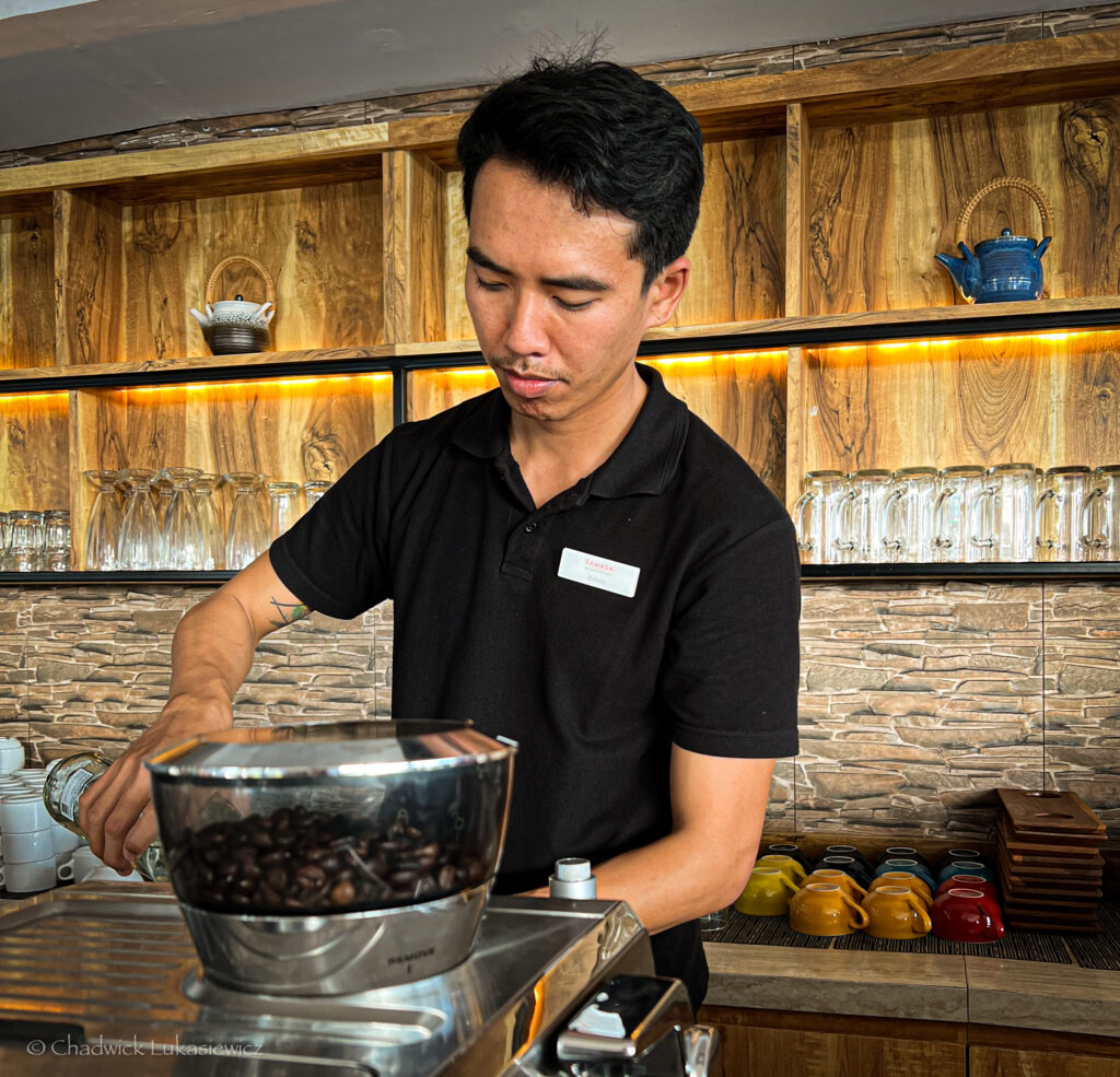 A bartender at the Ramada in Kuta, Bali, wearing a black polo shirt with a name tag, carefully pouring a liquid into a coffee grinder filled with dark roasted coffee beans. The bar features wooden shelves with glassware and decorative items in the background