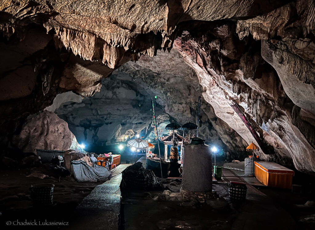 Inside a large, dimly lit limestone cave, an Indonesian temple is set up with decorative umbrellas, offerings, and small altars illuminated by spotlights. The cave’s jagged rock formations loom overhead, and the floor is lined with colorful checked cloths. The sacred atmosphere of the temple contrasts with the natural roughness of the cave, creating a serene yet dramatic scene.