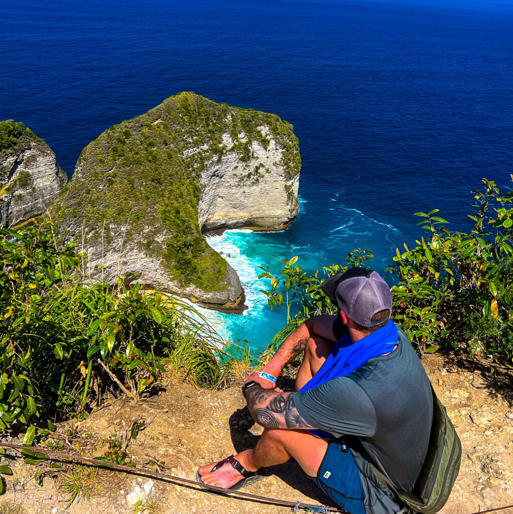 A person with a tattooed arm sits on the edge of a cliff overlooking the turquoise waters and iconic rock formations of Kelingking Beach on Nusa Penida, Bali. The rock formation below resembles the shape of a T-Rex head, surrounded by deep blue ocean waters. Lush greenery surrounds the cliffside, while the individual, wearing casual outdoor gear and sandals, gazes out at the breathtaking view. The scene captures a moment of contemplation and adventure amidst a striking natural landscape.