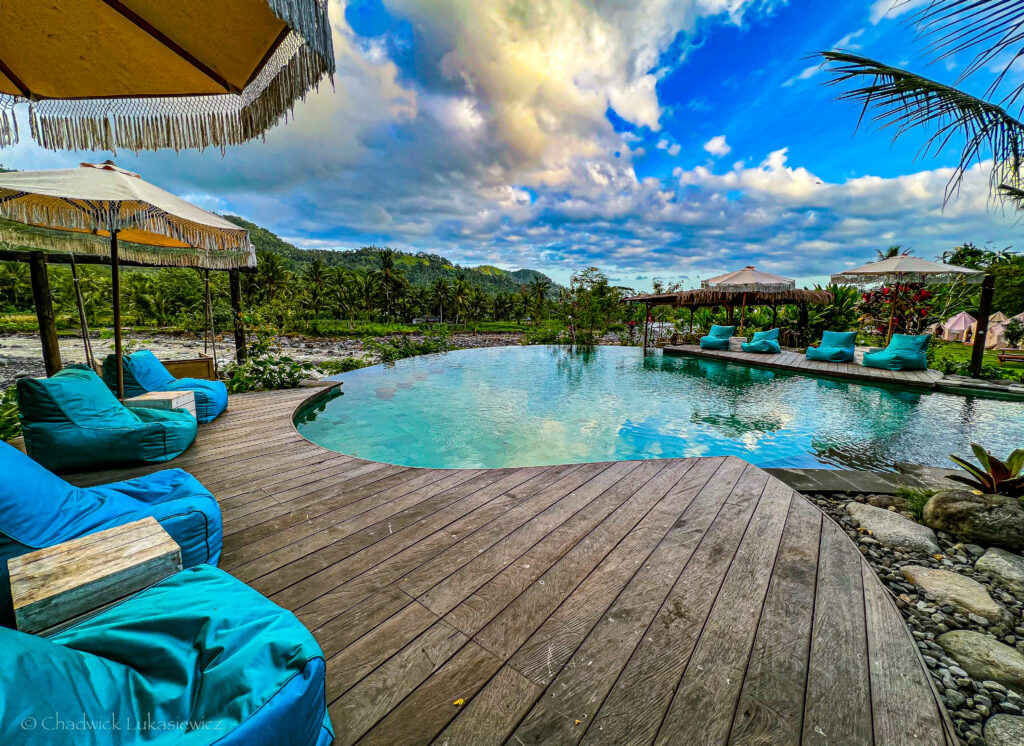 A serene poolside scene at a tropical resort, featuring a wooden deck with vibrant turquoise beanbag chairs and large, fringed parasols. The infinity pool reflects the partly cloudy sky, blending into the lush, green landscape of palm trees and hills in the distance. The calm ambiance evokes a peaceful, luxurious escape in nature.