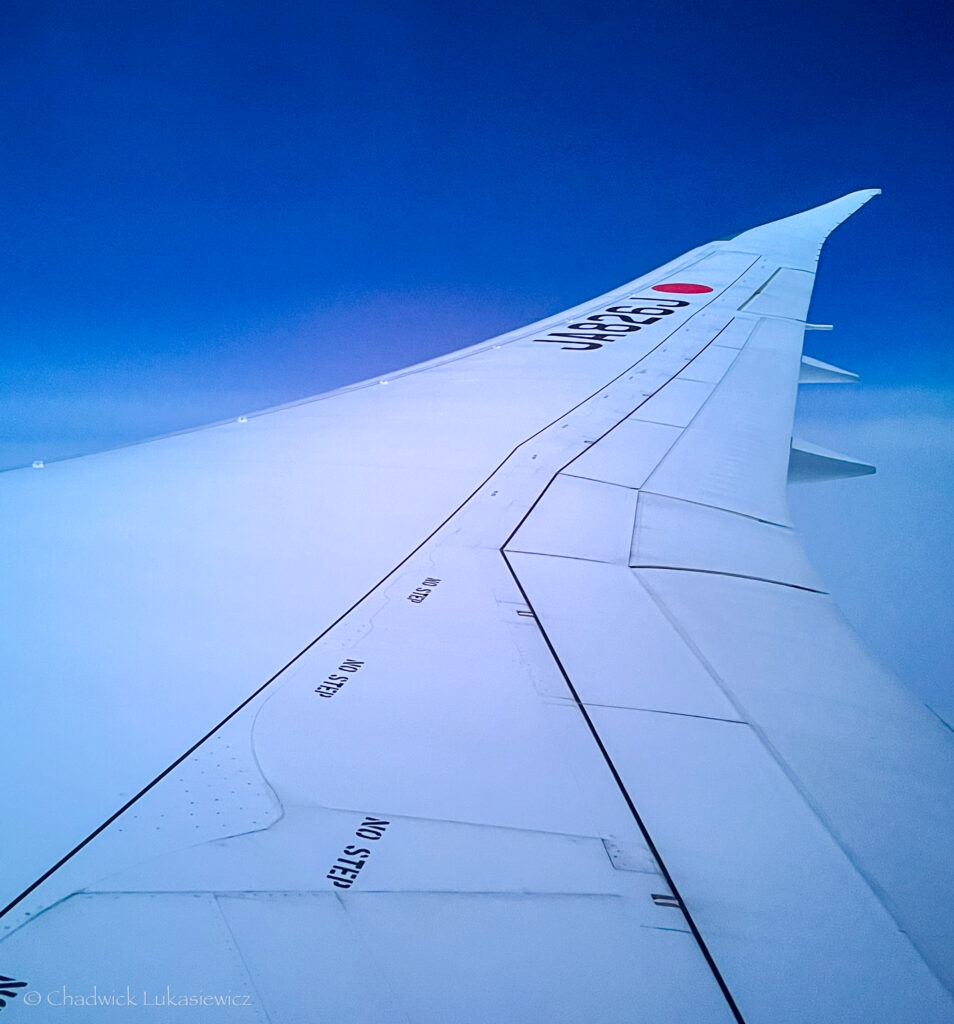 View from an airplane window showing the large wing of a Japan Airlines aircraft (JAL) against a clear blue sky. The white wing features markings like ‘NO STEP,’ and the iconic red circle of the airline’s logo is visible near the tip. The scene captures the peacefulness of flight at high altitude.