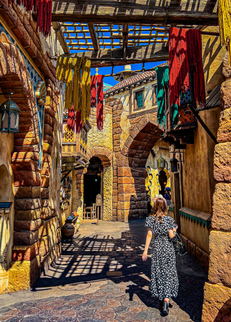 A woman in a black and white polka-dot dress walks through a sunlit alleyway with textured stone walls and arched doorways in a themed setting. Colorful fabric tassels hang from wooden beams above, casting intricate shadows on the stone path below. The architecture and decor evoke a Middle Eastern or Arabian-inspired atmosphere, with ornate lanterns and pottery visible along the sides. The bright blue sky contrasts with the warm tones of the surroundings, creating a vibrant and immersive scene.