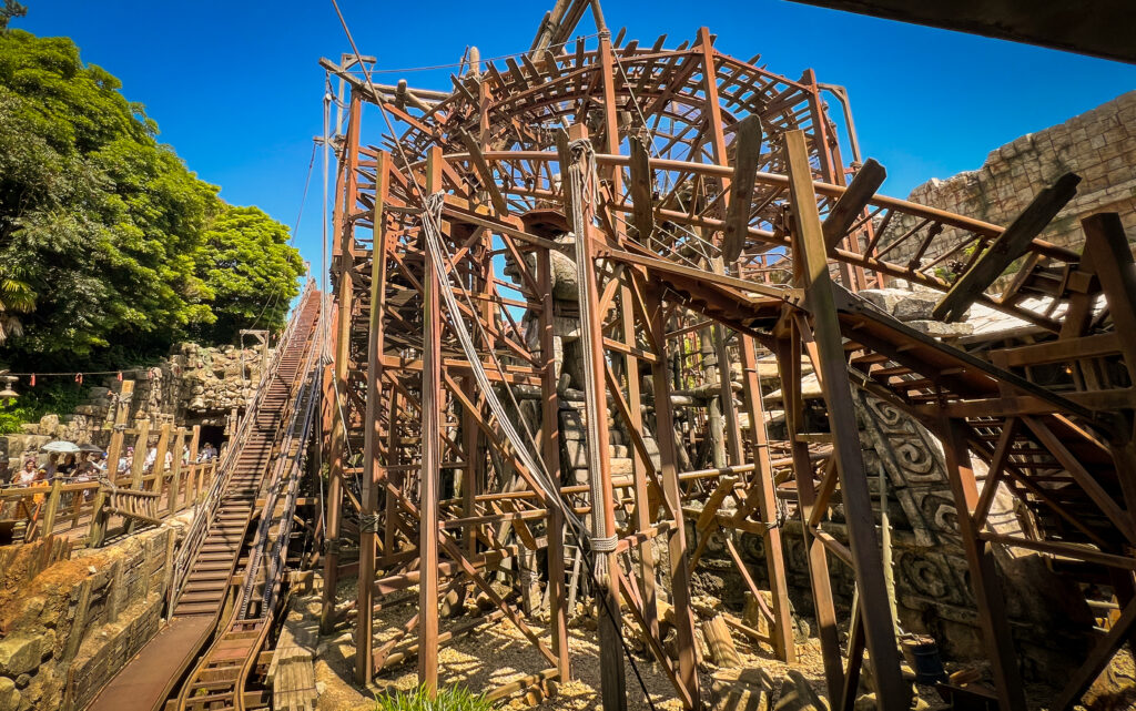 An intricate wooden and metal roller coaster structure at Tokyo DisneySea’s Raging Spirits ride. The ride’s design features steep tracks, wooden beams, and ropes, creating an adventurous atmosphere reminiscent of an ancient archaeological site. In the background, lush green trees and rock formations frame the scene, while a vibrant blue sky stretches overhead, adding to the thrill of the ride.