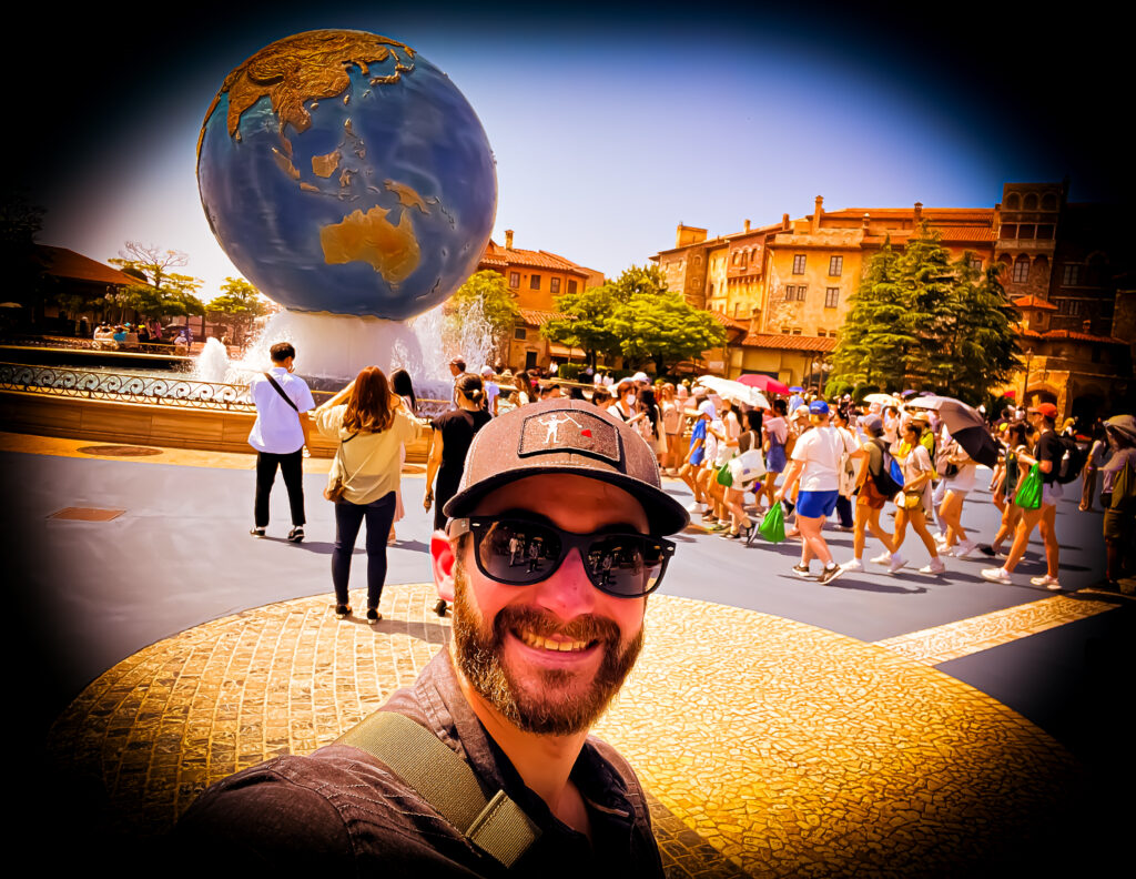 A man taking a selfie at Tokyo DisneySea in front of the iconic AquaSphere, a large globe fountain with continents detailed in gold. The man wears sunglasses, a dark cap with a logo, and smiles broadly at the camera. Behind him, visitors are walking around, some holding umbrellas for shade, and the background shows Mediterranean-style buildings bathed in bright sunlight. The vivid colors and cheerful atmosphere reflect the excitement of a theme park visit.