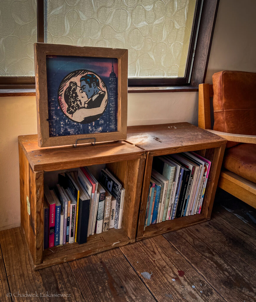 A cozy interior scene featuring a wooden shelf made from two crates, filled with books and magazines. On top of the shelf, a framed print depicts a stylized couple embracing against the backdrop of a city skyline with the Empire State Building. The warm sunlight filters through a textured window, casting soft shadows on the wooden floor and shelf. To the right, part of a brown leather chair is visible, adding to the comfortable and rustic ambiance of the space.