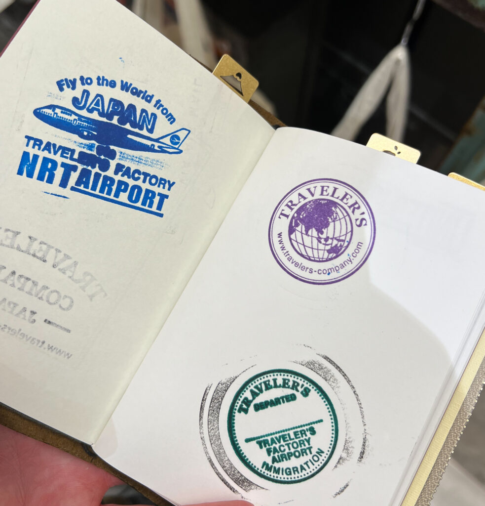 A Traveler’s Notebook opened to a page displaying three stamps from the Traveler’s Factory at Narita Airport. The top-left stamp is blue, featuring an airplane graphic and the text “Fly to the World from Japan, Traveler’s Factory NRT Airport.” The top-right stamp is purple, with the Traveler’s Company logo, showing a globe and website. The bottom stamp is green, resembling an immigration stamp, with the words “Traveler’s Factory Airport Immigration” and “Departed.” The notebook corners are held with brass clips, enhancing the travel-themed aesthetic.