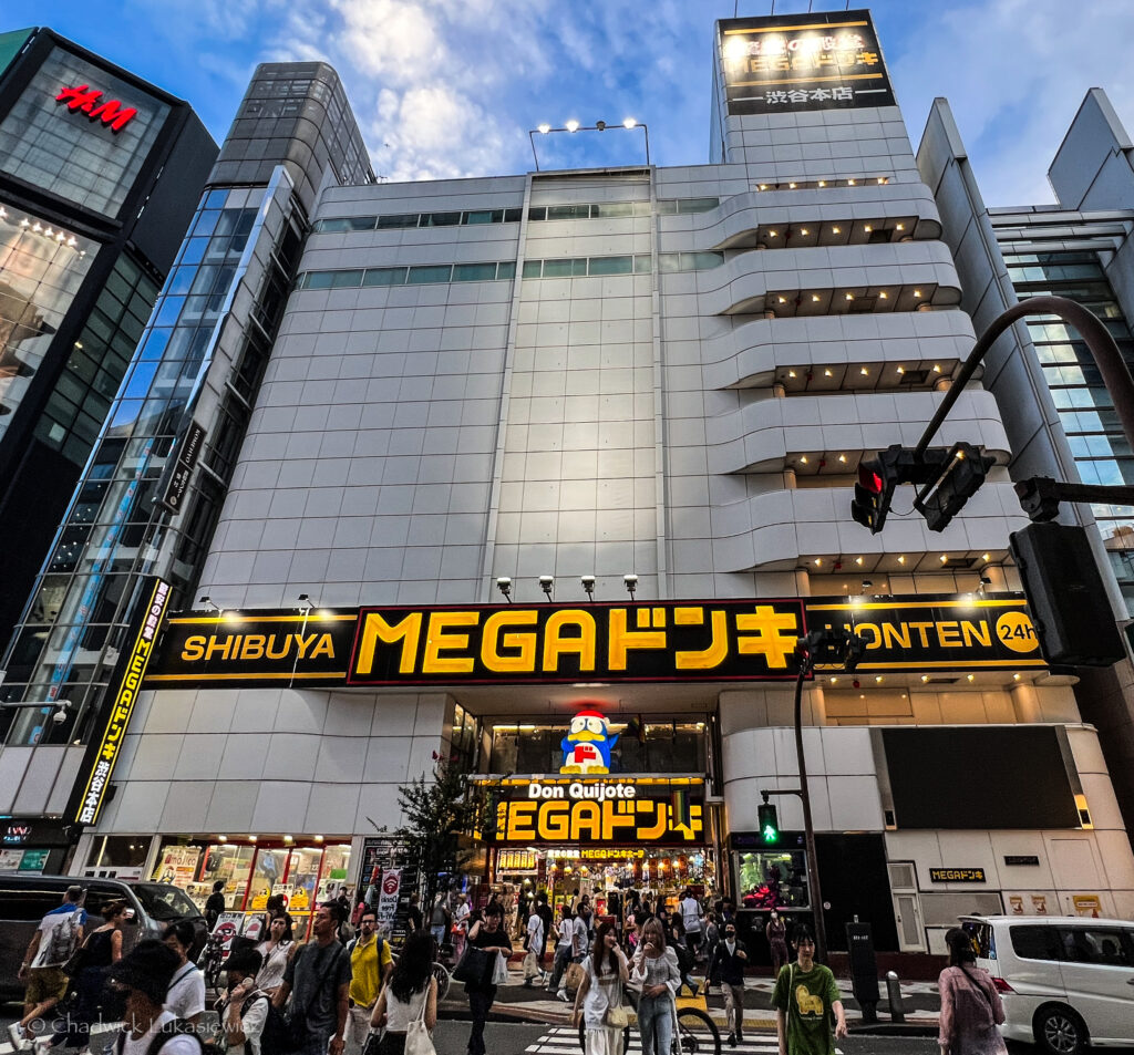 A bustling street scene in front of the Don Quijote MEGA store in Shibuya, Tokyo. The large, modern building features the store’s bright yellow ‘MEGA Don Quijote’ sign, prominently displayed above the entrance. Shoppers walk past the store’s entrance, and traffic lights and street signs are visible in the foreground. To the left, the ‘H&M’ store sign is partially visible on an adjacent tall building. The sky is partly cloudy as dusk begins to fall, casting a soft light on the scene.