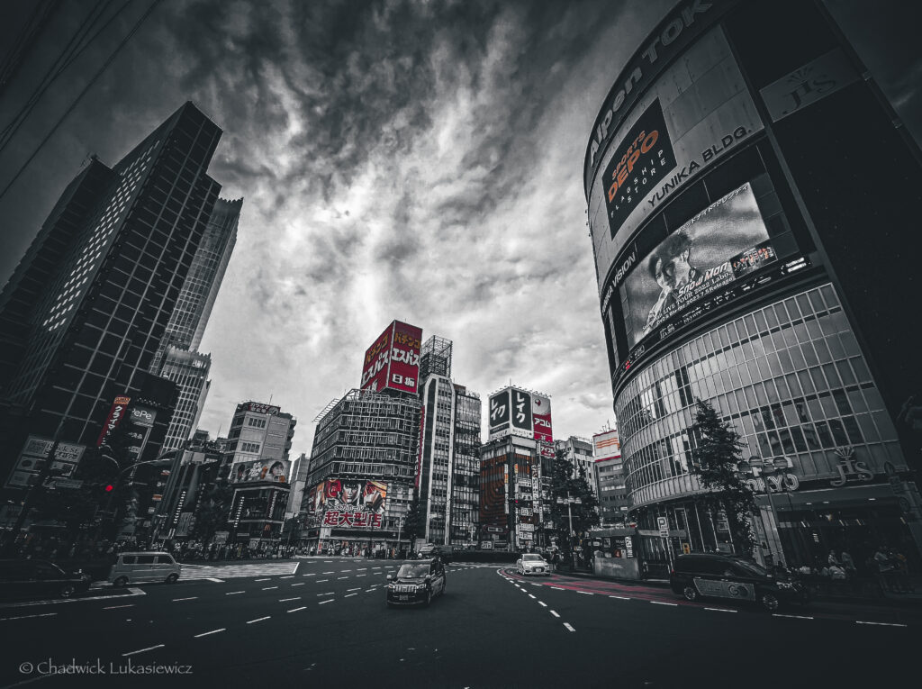 Black-and-white photo of a wide street intersection in Shinjuku, Tokyo, surrounded by tall buildings. Large digital billboards and advertisements, some in bright red, dominate the skyline. The scene captures a few cars driving along the road under an overcast sky, creating a moody and dramatic urban atmosphere.