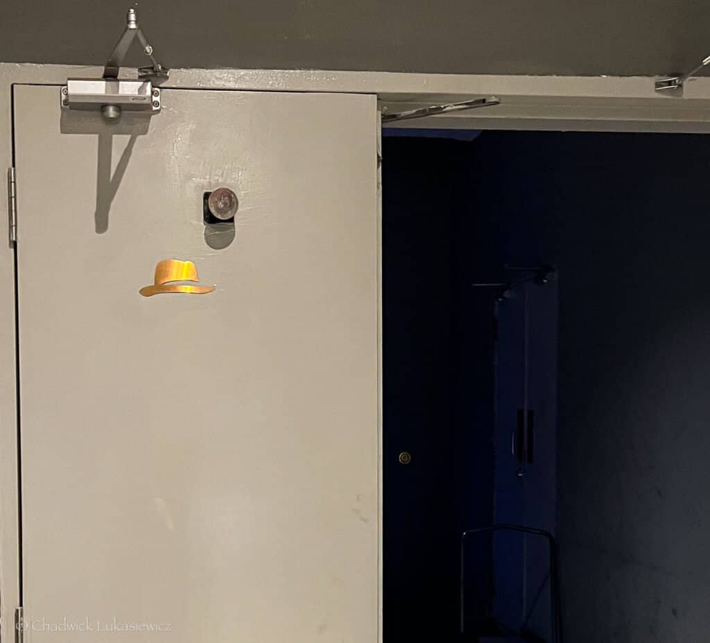 A plain door with a small, illuminated gold hat symbol, partially open to reveal a dimly lit room beyond, taken at Frank’s Bar in Kuala Lumpur.