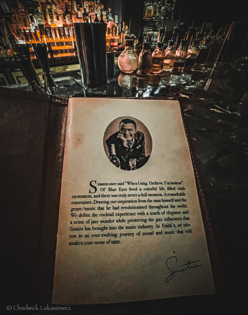 Dimly lit bar with an open menu featuring a portrait of Frank Sinatra and a quote attributed to him. The menu highlights Sinatra’s influence on jazz and cocktails. In the background, an array of glass bottles and bartending equipment can be seen on the bar, adding to the vintage and sophisticated ambiance of the space