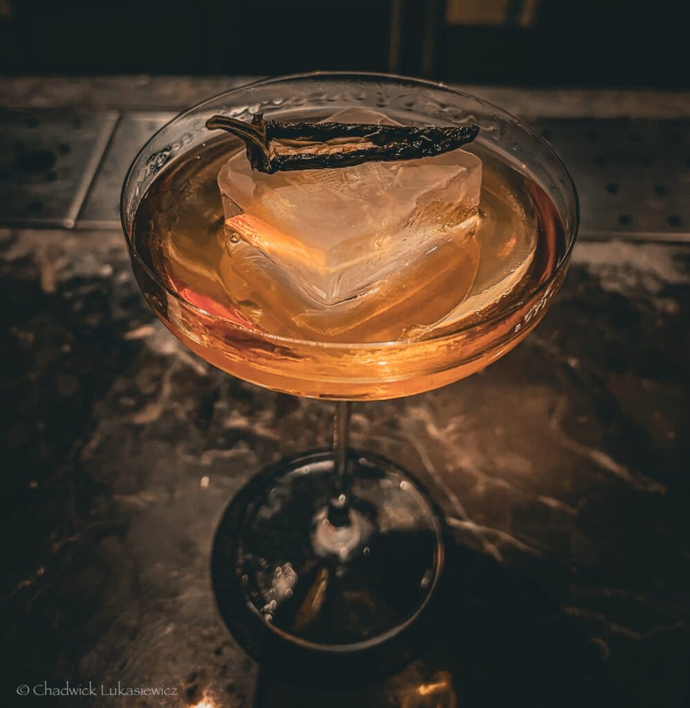 A close-up of a cocktail in a coupe glass with a large, clear ice cube and a dried garnish on top, served on a dimly lit marble bar at Frank’s Bar in Kuala Lumpur.