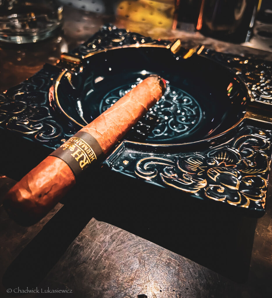 A single cigar rests on an ornate black ashtray with intricate gold detailing. The cigar has a visible label reading ‘Herbalano.’ The scene is dimly lit, with a warm, moody ambiance reflecting off the metallic surface of the table beneath the ashtray.