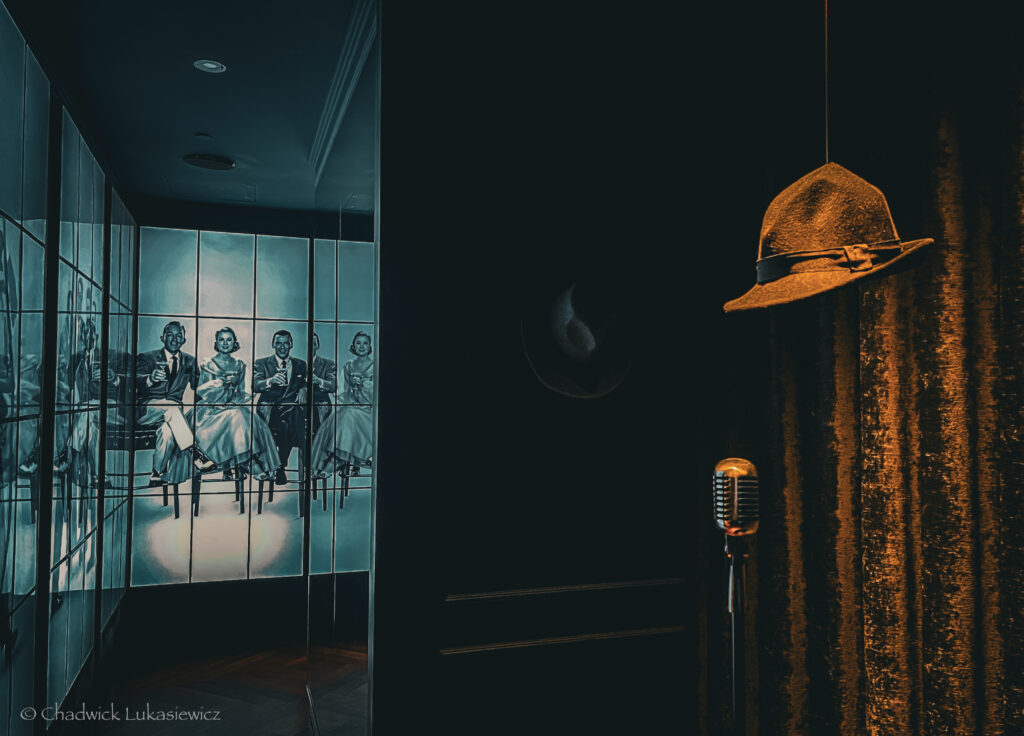 A dimly lit lounge scene featuring a classic vintage microphone in the foreground, with a fedora hat suspended above it. In the background, mirrored panels reflect a black-and-white mural of elegantly dressed people seated in a theater-style setting, creating a nostalgic and moody ambiance