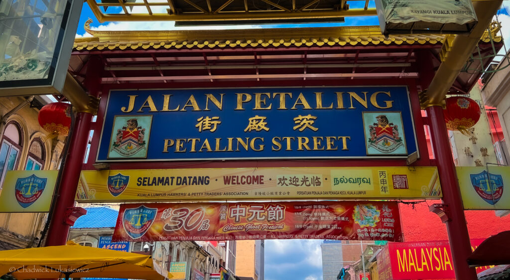 A close-up of the entrance arch to Petaling Street in Kuala Lumpur, Malaysia, showcasing a vibrant blue and gold sign reading “Jalan Petaling” in both English and Chinese. The sign is framed by decorative red pillars and traditional gold accents, with various smaller signs below welcoming visitors in multiple languages. A festive banner for the “Chinese Ghost Festival” hangs underneath. Bright red lanterns and buildings can be seen in the background, adding to the lively atmosphere.