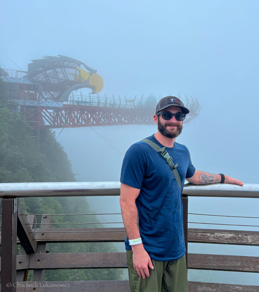 A man wearing a blue shirt, green pants, and sunglasses stands on a misty observation deck in Langkawi, Malaysia. Behind him, a fog-covered bridge with an eagle-shaped structure emerges from the mist, blending into the surrounding greenery