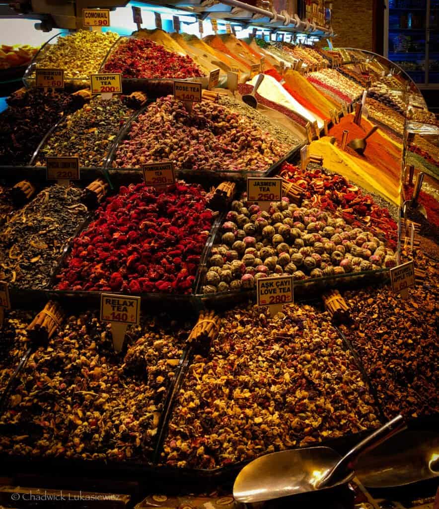 A vibrant display of various teas and spices arranged in piles at a market. The colorful assortment includes hibiscus tea, rose tea, love tea, and many others, with signs indicating their names and prices. The mounds of dried herbs, flowers, and spices create a rich tapestry of colors, ranging from deep reds and pinks to yellows and greens. Small wooden scoops rest atop the piles, and a large metal scoop is visible in the foreground, ready for serving. The market’s warm lighting enhances the cozy and inviting atmosphere.