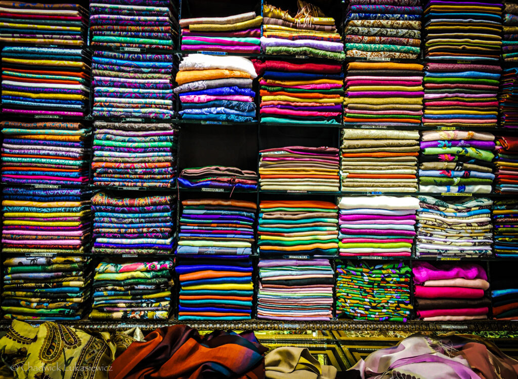 “A vibrant display of neatly folded fabrics arranged on shelves in a shop. The fabrics come in a variety of colors, patterns, and textures, including intricate paisley designs, florals, and solid colors. At the bottom, a few colorful pieces are loosely draped, adding to the vibrant array. The shelves are stacked floor to ceiling, showcasing a rich assortment of textiles.