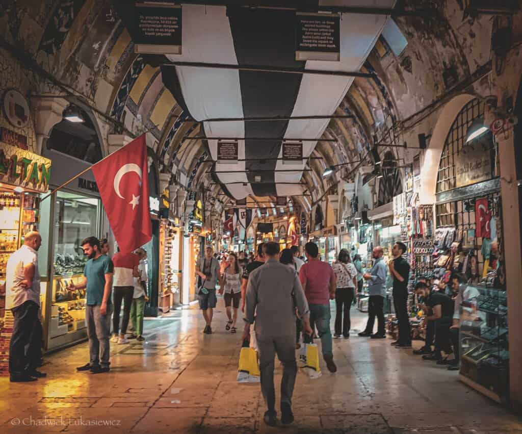 Busy interior of the Grand Bazaar in Istanbul, Turkey, with a Turkish flag prominently hanging on the left. Shoppers walk down the corridor, passing by various small shops selling jewelry, souvenirs, and local goods. The ceiling is adorned with alternating black and white cloth panels, giving a traditional yet vibrant atmosphere.