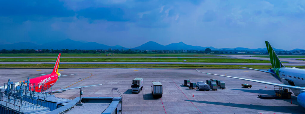 The image depicts an airport runway with several planes visible. The foreground shows the tail sections of two commercial planes from VietJet Air and another airline. There are also airport service vehicles on the tarmac, such as luggage carts and transport trucks. The background features a grassy runway with distant mountains under a hazy, cloudy sky. The scene conveys a typical airport atmosphere with calm activity on the ground.