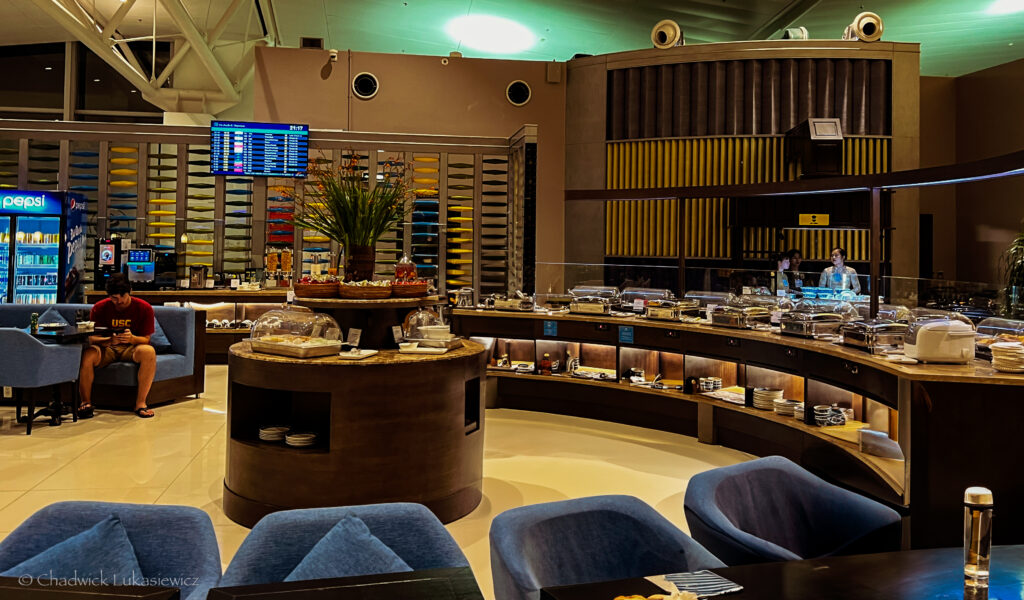 A spacious airport lounge with a circular buffet area at its center, offering various food items covered under glass domes and chafing dishes. A person wearing a red USC shirt is seated on a blue chair, looking at their phone while dining. The lounge features a Pepsi vending machine, a beverage station, and flight information displayed on a digital screen above. The decor includes wood-paneled accents and colorful design elements, creating a modern and comfortable atmosphere.