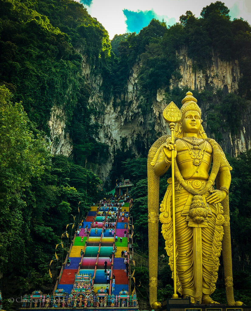 A towering golden statue of Lord Murugan stands in front of the Batu Caves in Kuala Lumpur, Malaysia. Behind the statue, a long, vibrant staircase painted in a rainbow of colors leads up to the cave entrance, with visitors climbing the steps. The surrounding cliffs are covered in lush greenery, creating a dramatic natural backdrop. The sky is partly cloudy, with sunlight illuminating the scene.