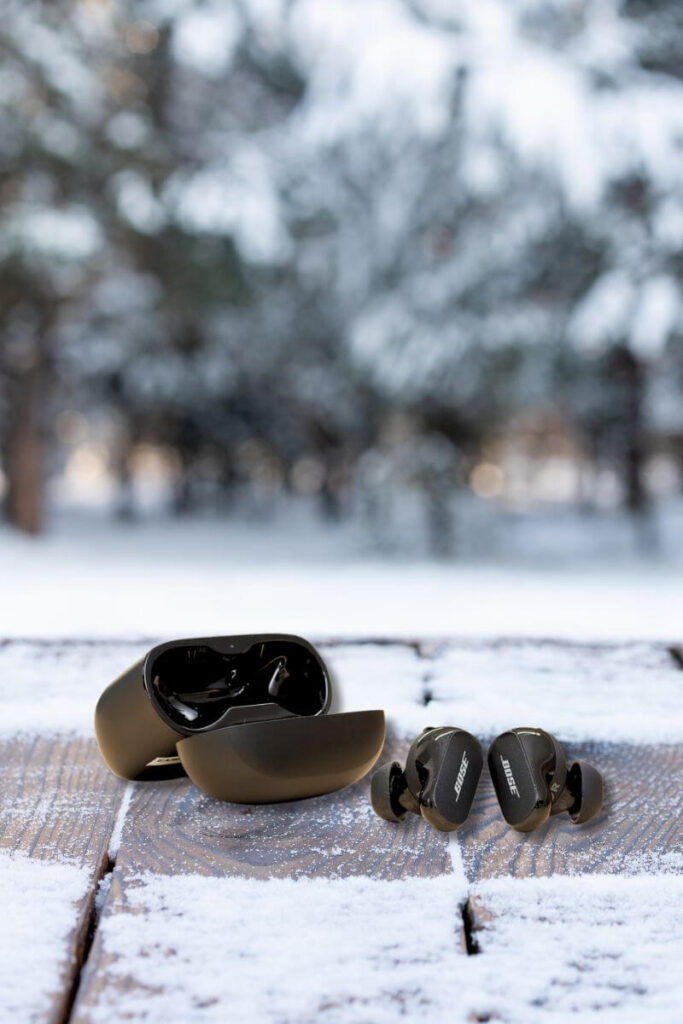 A pair of black Bose QuietComfort earbuds placed on a snow-dusted wooden table outdoors, next to their matching open charging case. The blurred background features snowy trees.