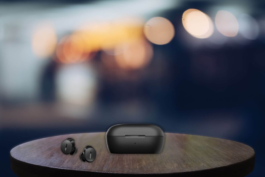 Black Bose QuietComfort II earbuds and their matching charging case displayed on a round wooden table, with a softly blurred background featuring warm bokeh lights and a dark ambiance.