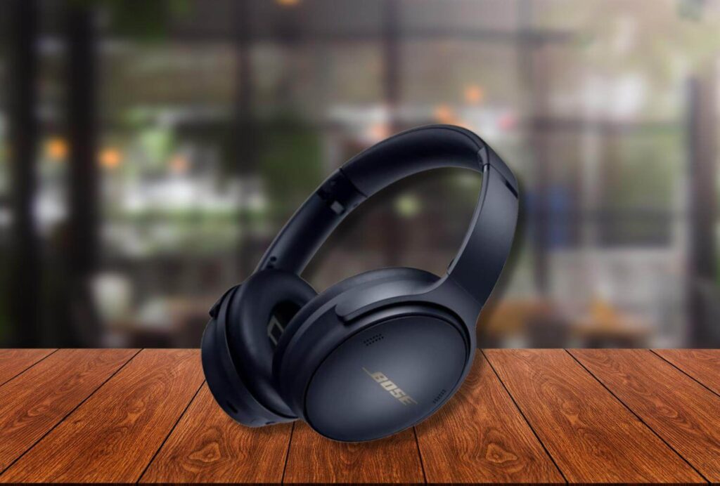 A pair of black Bose QuietComfort 45 headphones placed on a wooden table, with a softly blurred café interior in the background featuring warm lighting and greenery