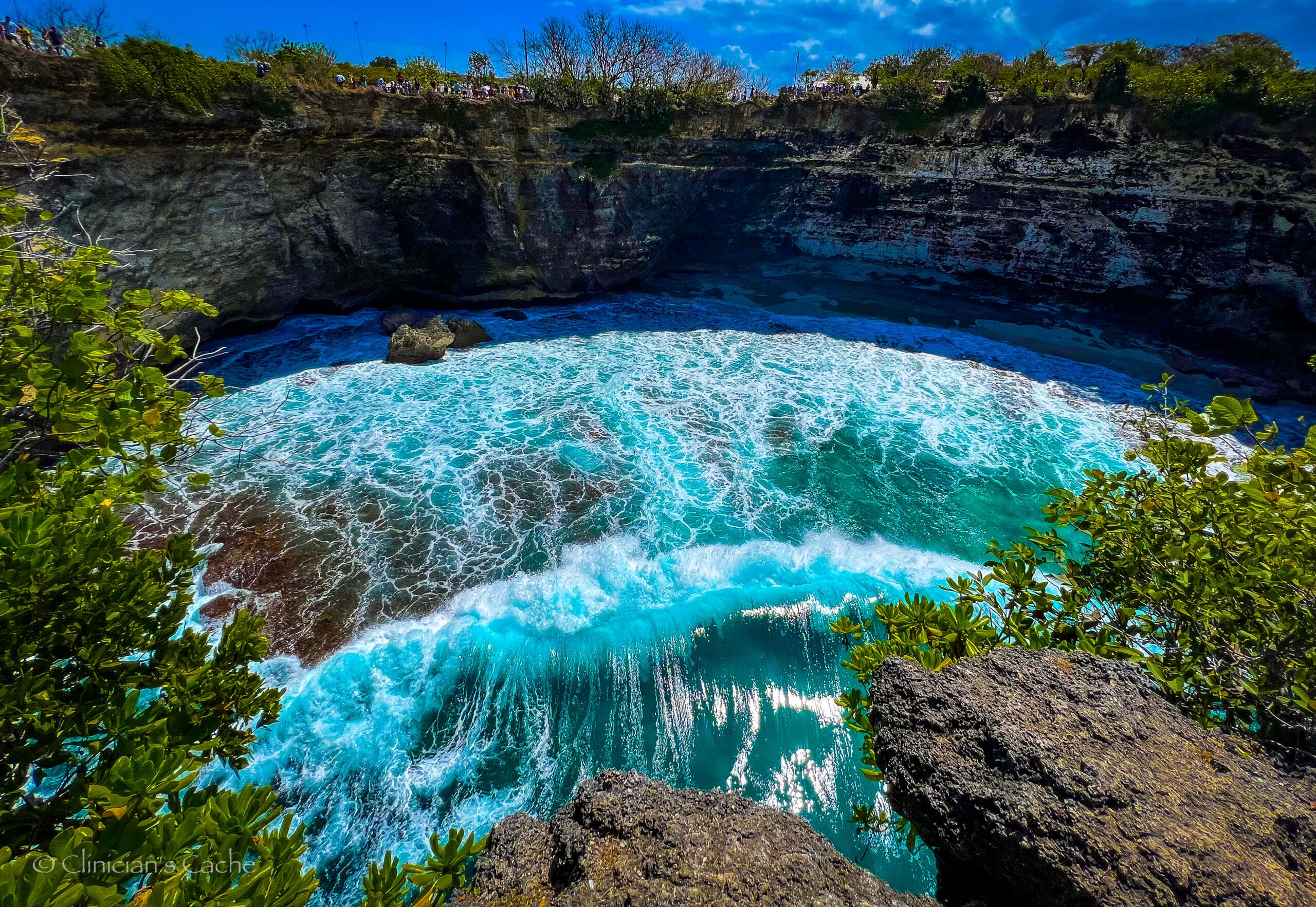 Turquoise waves crash into a circular cove surrounded by rocky cliffs at Broken Beach, Nusa Penida, Bali, with tourists watching from the cliff edge above.