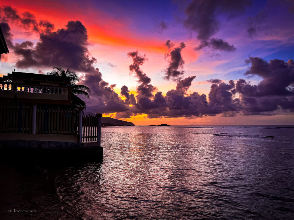 Vibrant sunset over calm ocean waters, with dramatic clouds and a house silhouetted along the shoreline in Calibishie, Dominica