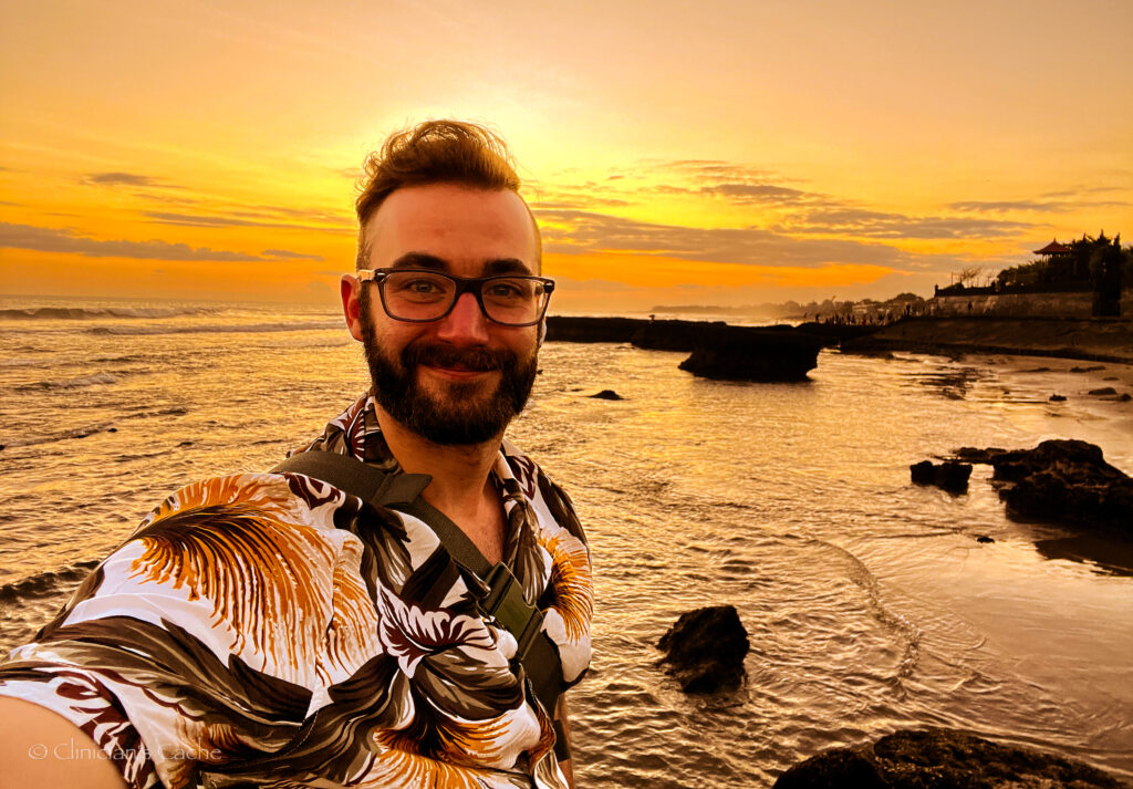 Man with beard and glasses taking a selfie at sunset on a beach in Bali, Indonesia, with calm waves and a golden sky in the background.