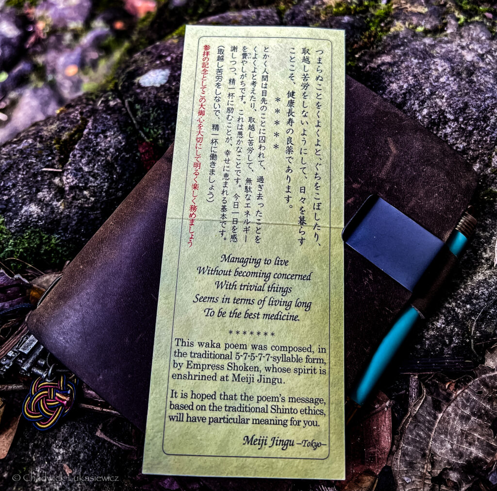 A rectangular card with text in Japanese and English rests on a weathered stone surface surrounded by moss and leaves. The card contains a waka poem, translated in English as: “Managing to live / Without becoming concerned / With trivial things / Seems in terms of living long / To be the best medicine.” Below the poem, additional English text explains that the waka poem is composed in the traditional 5-7-5-7-7 syllable form by Empress Shoken, whose spirit is enshrined at Meiji Jingu, Tokyo. It expresses a wish for the poem’s message, rooted in Shinto ethics, to hold personal significance for the reader. Beside the card is a teal and black pen and a leather notebook with a decorative knot attached to it. The vibrant green and earthy tones of the surroundings add a serene and contemplative ambiance to the scene