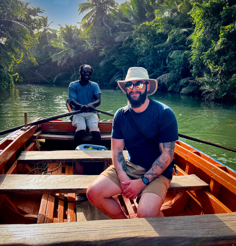 A man wearing sunglasses and a sunhat sits smiling on a wooden boat as another man rows behind him on a lush, green river. Palm trees and dense vegetation surround the water, creating a serene tropical atmosphere.