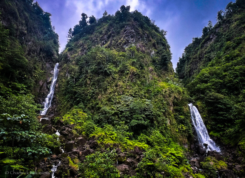 Scenic view of Trafalgar Falls in Dominica, featuring two towering waterfalls cascading down lush, green mountainsides. The twin waterfalls are surrounded by dense tropical vegetation, with rocks scattered at the base of the falls. The overcast sky adds a moody ambiance to the dramatic natural landscape.