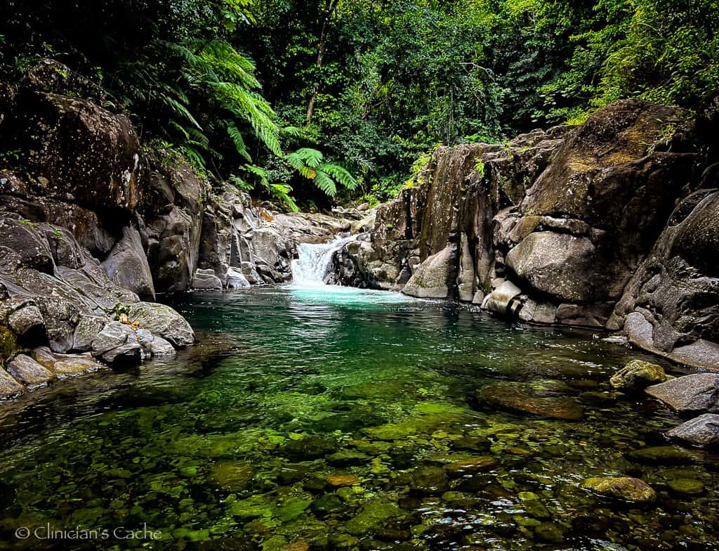 A serene natural pool with clear emerald green water surrounded by rocky cliffs and lush tropical vegetation. A small waterfall gently cascades into the pool, creating a peaceful, secluded atmosphere in the midst of the forest.