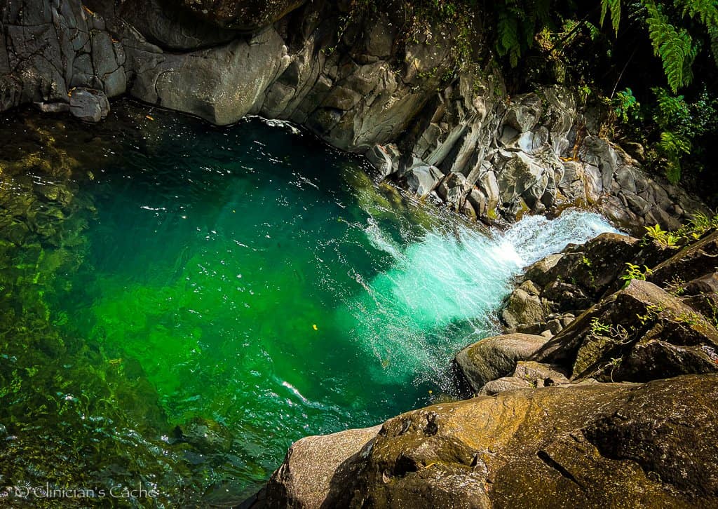 “A crystal-clear natural pool with vibrant green and turquoise hues surrounded by rocky cliffs and lush greenery. Water flows gently into the pool from a small cascade, creating ripples across the serene surface.