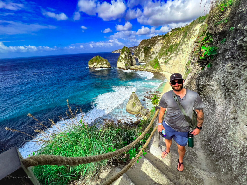 Man standing on a steep path with a rope railing overlooking the turquoise waters and rocky cliffs of Diamond Beach, Nusa Penida, Indonesia.