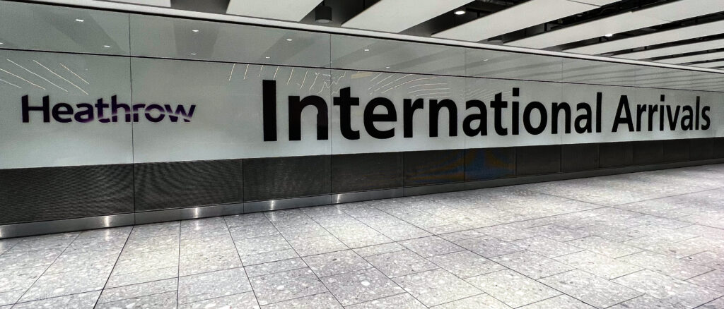 Sign at Heathrow Airport showing ‘Heathrow International Arrivals’ in bold black letters on a white wall with a modern design. The polished floor and ceiling lights reflect the sleek, clean look of the arrivals area.