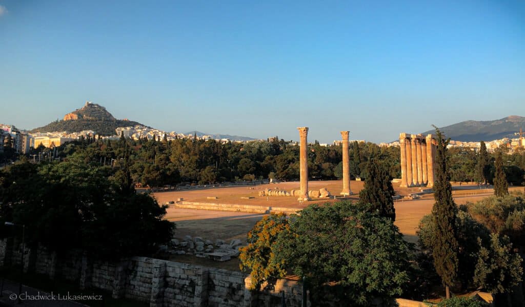 Ruins of the Temple of Olympian Zeus in Athens, Greece, with several standing columns, surrounded by greenery, and Mount Lycabettus in the background under a clear sky