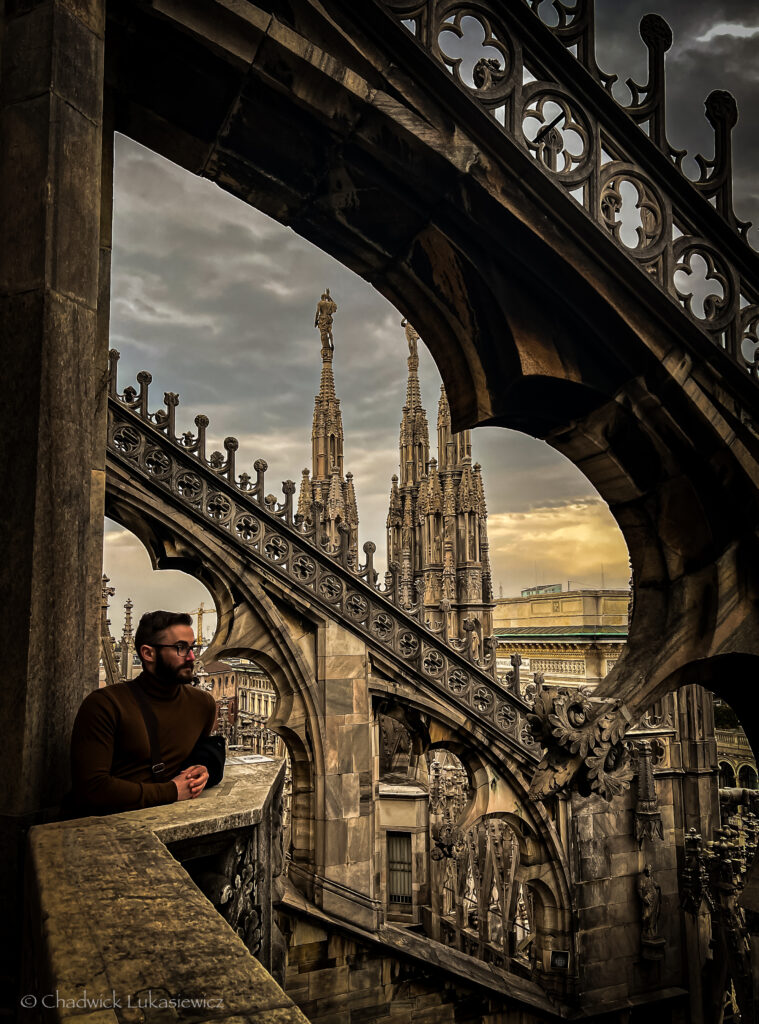 A man standing on a balcony of the Duomo di Milano, surrounded by intricate Gothic architecture. Arched stone structures frame the scene, while ornate spires rise into a moody, overcast sky. The man, dressed in a dark turtleneck, gazes thoughtfully into the distance. The majestic details of the cathedral’s design create a dramatic and historic atmosphere.