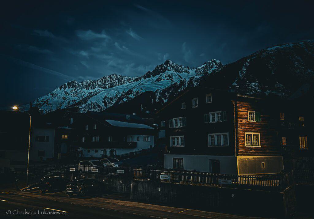 A serene winter night in Sedrun, Switzerland, with snow-covered mountains towering in the background, illuminated by the moonlight. Dark clouds add a moody atmosphere. In the foreground, warmly lit houses and parked cars create a cozy contrast against the chilly, mountainous landscape.